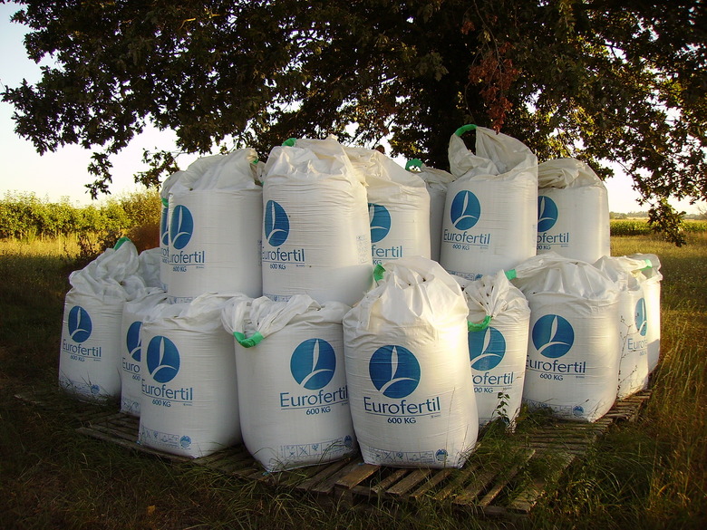 A group of large white bags of fertilizer stacked near a tree.