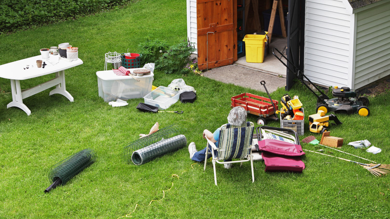 A woman sits near a garden shed with tools and other clutter scattered about.
