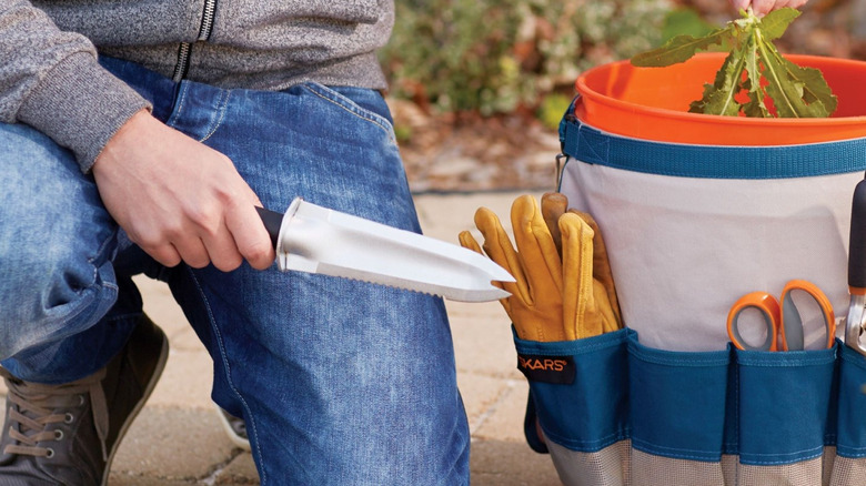 A gardener wearing a blue jeans holds the Fiskars garden knife next to a tool box