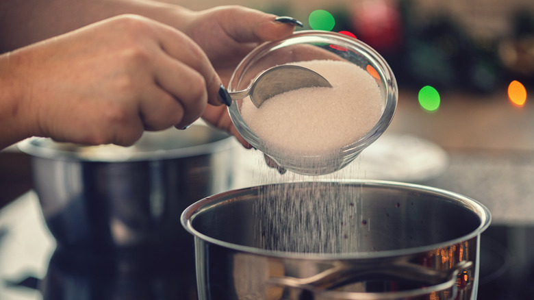 someone with black nails scooping sugar into a boiling pot of water