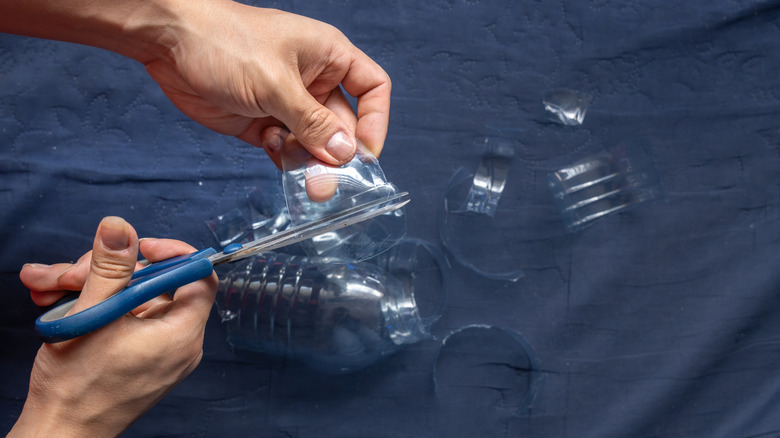 A close-up of hands using blue-handled scissors to cut strips from a clear plastic bottle