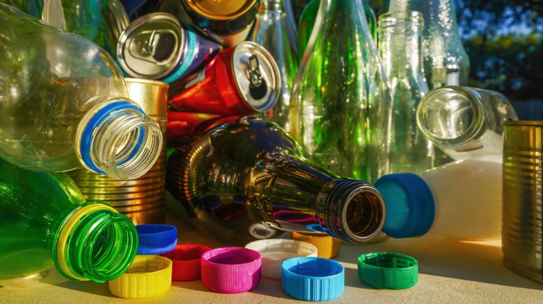 A colorful pile of recyclable waste, including plastic bottles, glass bottles, metal cans, and various brightly colored plastic caps scattered on a flat surface.