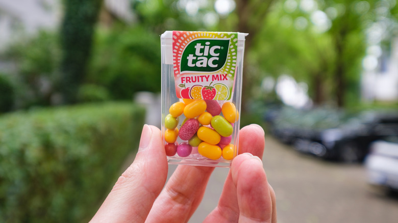 A hand is holding up a plastic container of Tic Tacs, filled with bright multicolored oval candies.