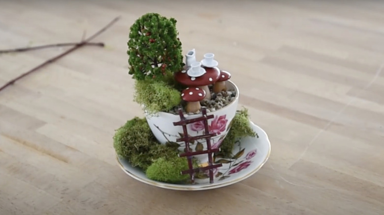 A white and pink tea cup and saucer is repurposed as a fairy garden with moss, a miniature tree, red-capped mushrooms, and a tiny tea set.