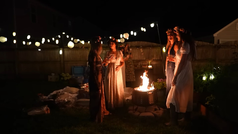 A group of women gather around a fire pit with fairy lights in the background at a "Practical Magic" themed party.