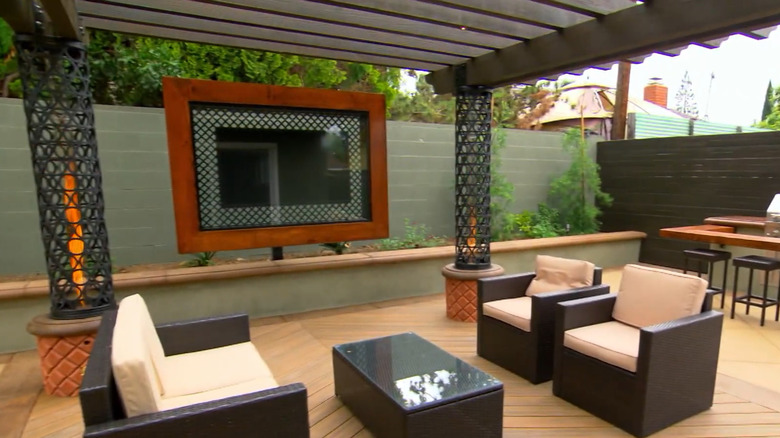 A television is mounted under a pergola to create an outdoor living space with chairs and a table.