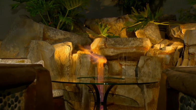 A small flame glows from a gas fireplace and water streams from a waterfall in the background.