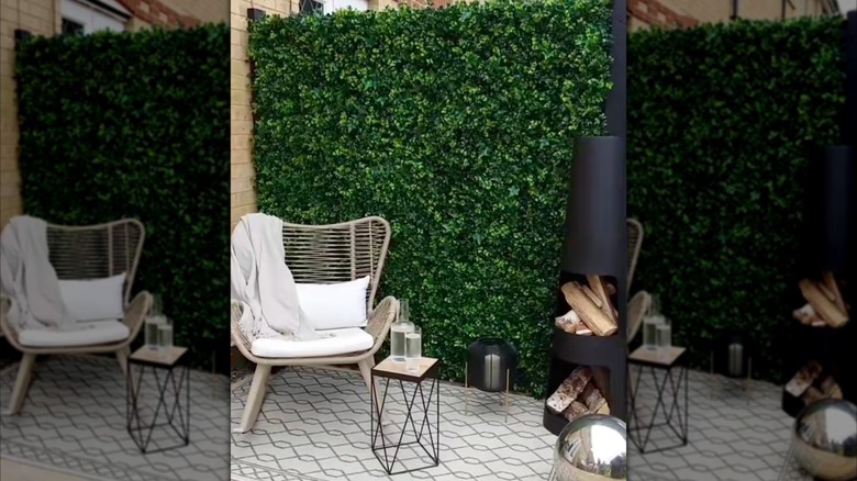A backyard patio is decorated with a gray chair, black metal side table, tall narrow wood-burning stove, silver gazing ball, with an artificial greenery wall in the background.