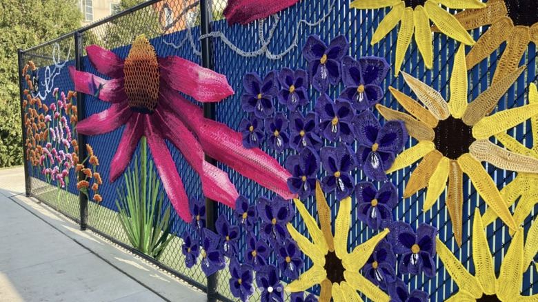Purple, yellow, pink, and orange flowers created from yarn are placed on a black chain link fence.