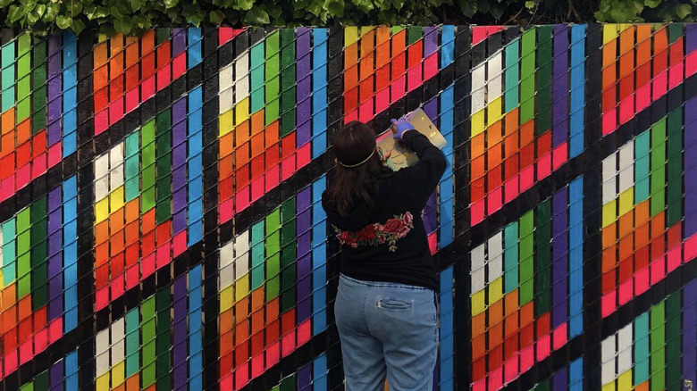 A woman with a black sweatshirt and blue jeans is standing in front of a chain link fence with colorful painted slats.