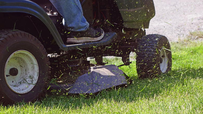 Closeup of the wheels of a riding mower in motion with the driver's tennis shoe and blue jeans visible