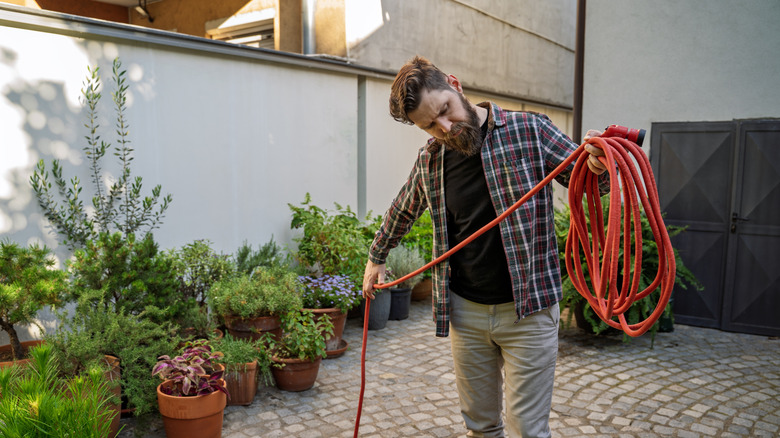 A bearded man in a plaid flannel shirt winding up an orange garden hose