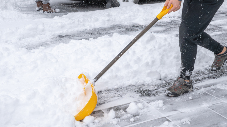 A person wearing black pants uses a yellow shovel to push snow