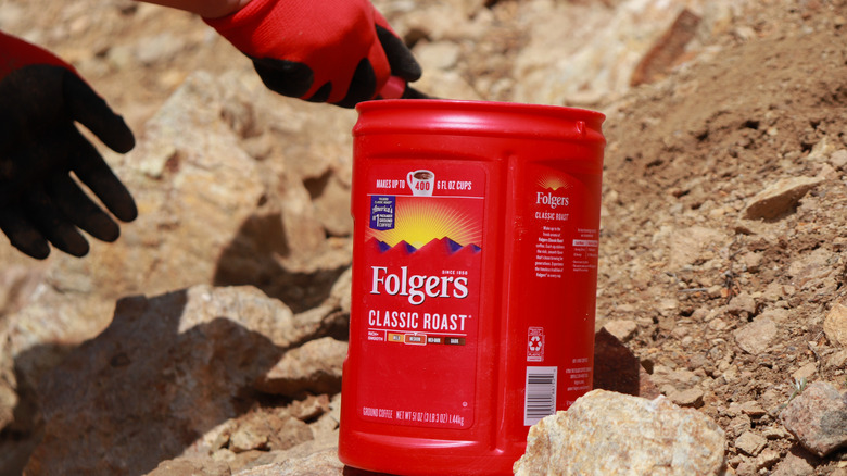 Someone setting a folgers coffee canister into the rocks while wearing gloves