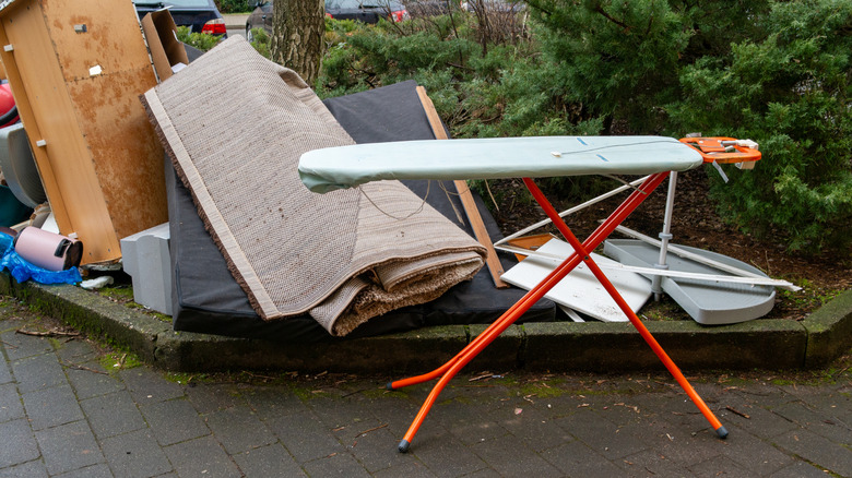 An orange and light blue ironing board is set up next to a pile of garbage.
