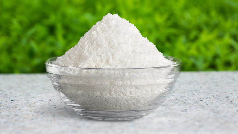 A clear glass bowl filled with white Epsom salt crystals, placed on a gray surface with greenery in the background.