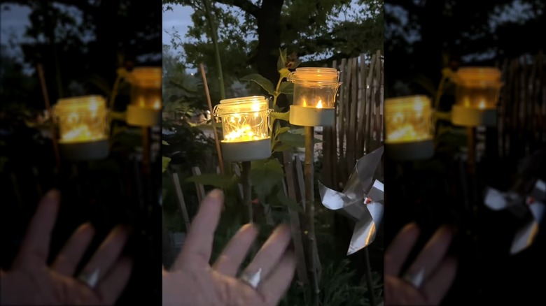 A hand gestures towards DIY garden lights made from old tuna cans, sticks, and mason jars.