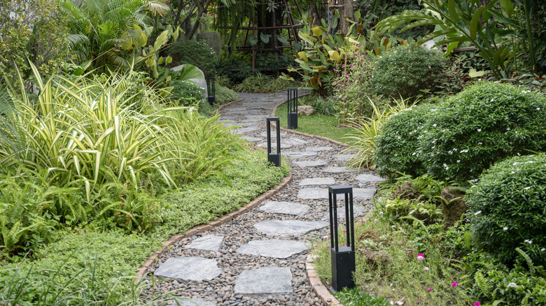A winding pathway of large pavers and small stones is lined with lights, grasses, ferns, and other shrubs.