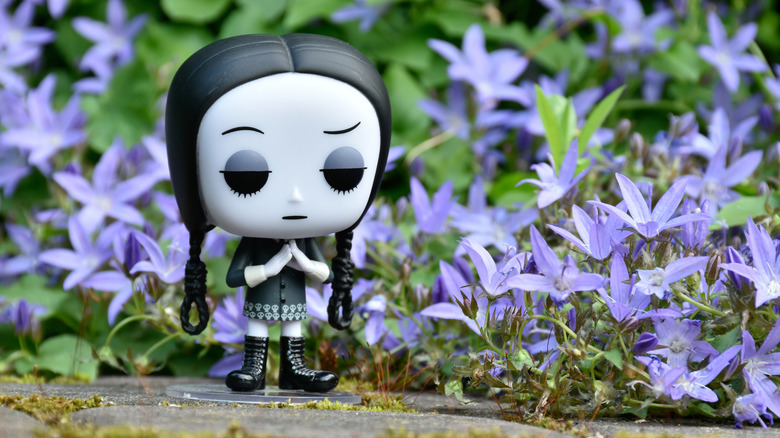 Pop vinyl figure of Wednesday stands in front of purple flowers.