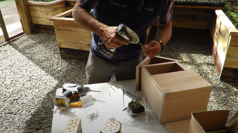 A man holds a drill over a wooden box with a roof on it.