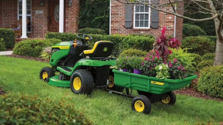 John Deere S100 lawn mower sits on lawn with trailer attached.