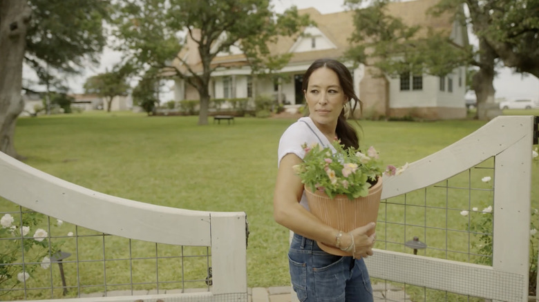 Joanna Gaines is holding a pot of flowers in a garden while opening a white and wire fence.