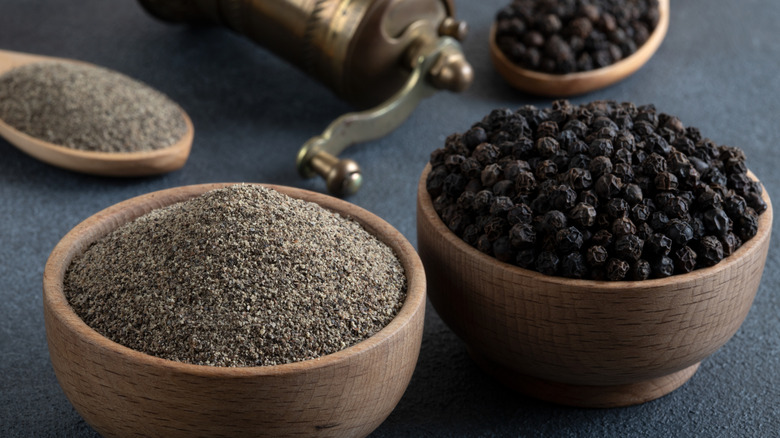 Wooden bowls of ground pepper and peppercorns are in the foreground, with wooden spoons holding ground pepper and peppercorns are in the background.
