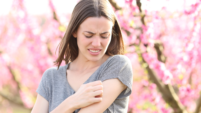 A white woman standing in front of a cherry blossom tree looks annoyed and pained as she scratches her arm.