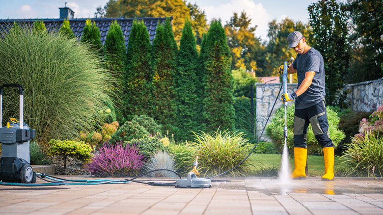 A man wearing yellow boots and gloves pressure-washing a patio in a landscaped backyard garden