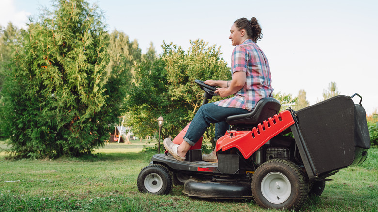 A woman riding a red riding lawn mower across a yard with trees in the background.