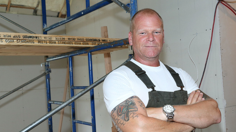 HGTV star Mike Holmes stands with his arms crossed while wooden planks and building equipment sit behind him.
