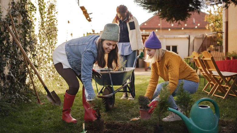 Three women work together in a garden with shovels and a wheelbarrow.