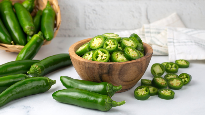 Chopped jalapenos in a bowl, next to several whole jalapenos on a kitchen counter.