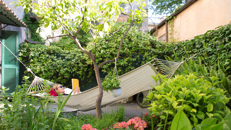 A woman relaxing in a hammock beneath greenery and tropical plants in a backyard garden.
