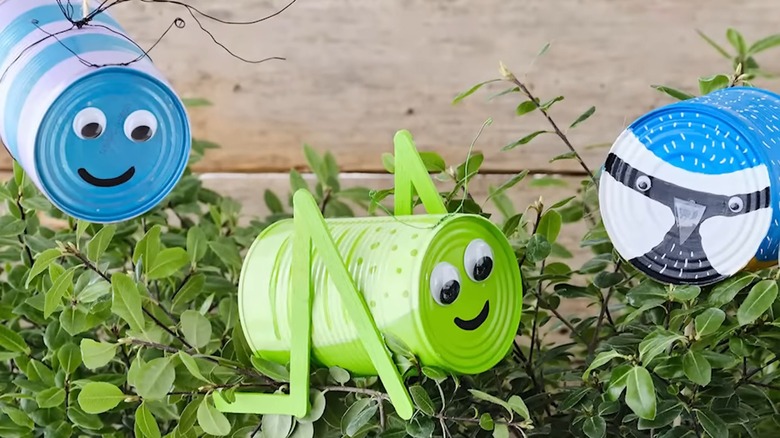 Soup cans painted like bugs and a bird decorate a garden.