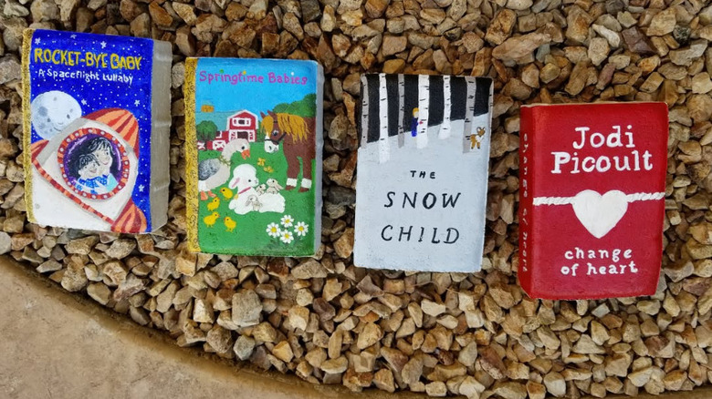 Four bricks painted like books sit in the hardscaping gravel of an outside garden.