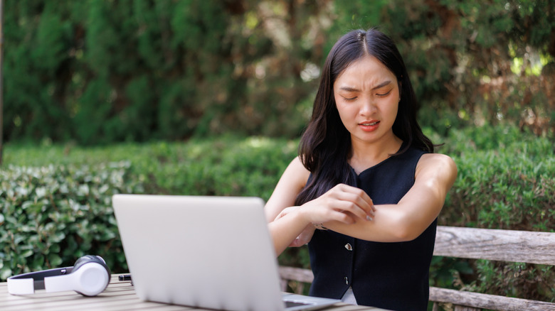 A young woman with dark hair scratching her arm in irritation while working outdoors with a laptop