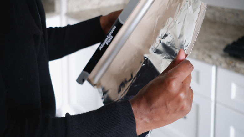 A pair of hands is pulling aluminum foil out of its narrow packaging.