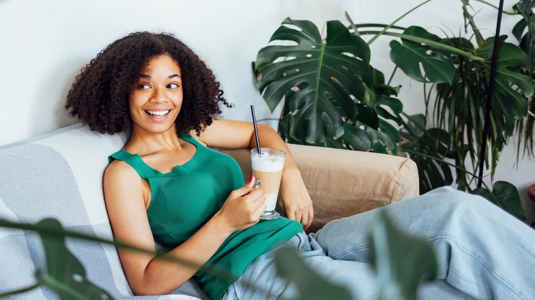 Woman with iced coffee sits near her monstera plant.