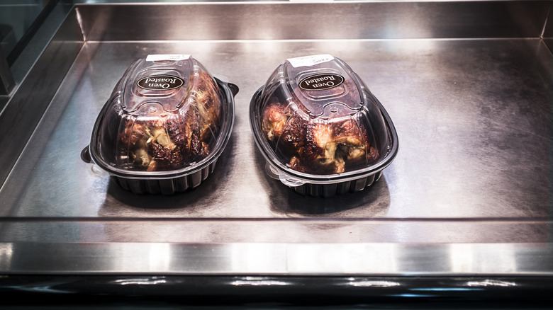 Two rotisserie chickens in a plastic container under a heat lamp on a metal serving surface.