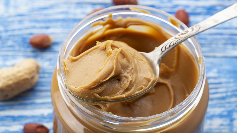 A spoonful of peanut butter sitting over a jar on a blue table with scattered peanuts.