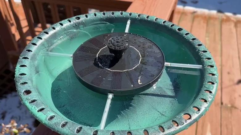 A birdbath with a fountain is clamped onto a deck railing.