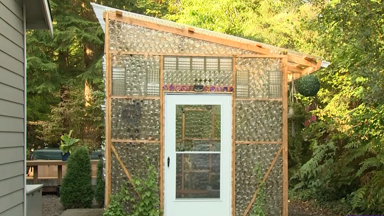 A greenhouse with walls made of pickle jars is in a garden next to a house.