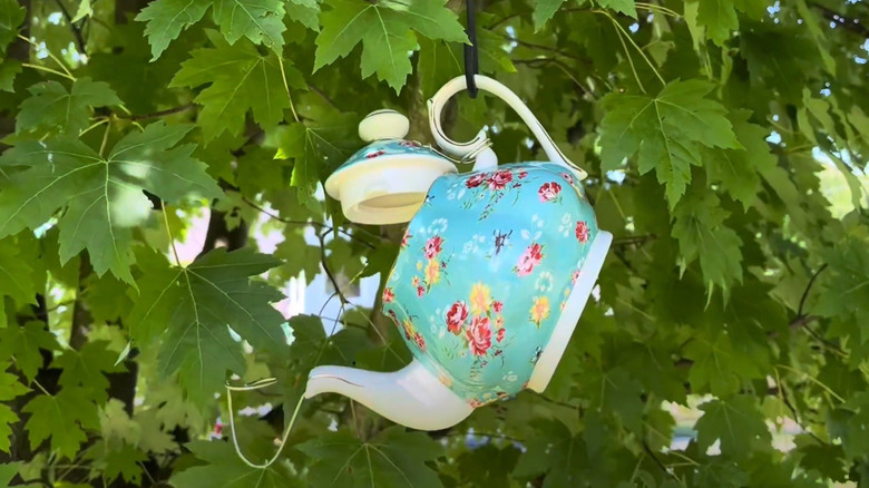 A teapot made to be a bird house hangs from a tree.
