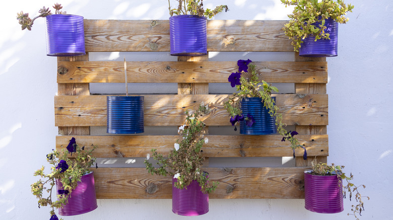 Painted tin cans holding flowers are nailed to a wooden pallet.