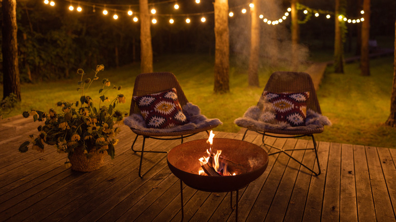 Fire burns in a metal firepit on a deck near two chairs.