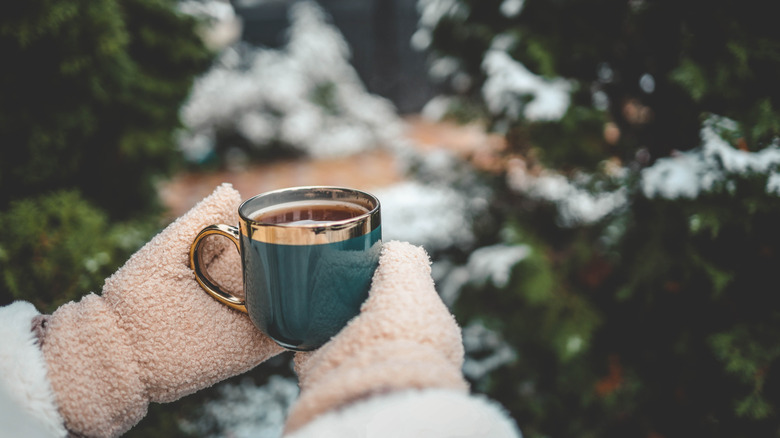 Hands covered in mittens hold a mug of tea outside in the winter.