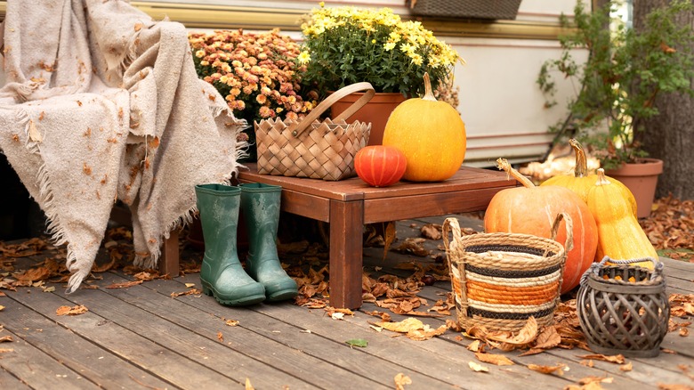 Wooden armchair with plaid near outside caravan trailer decorated for fall. RV house porch with garden furniture for relax nature. Interior cozy yard with rubber boots, flowers and pumpkins in garden.