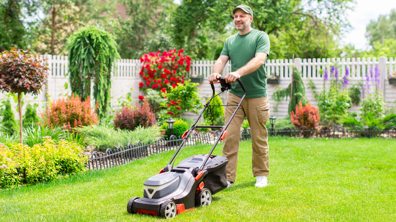 A man in a green shirt and brown pants mows his lawn with a push mower.